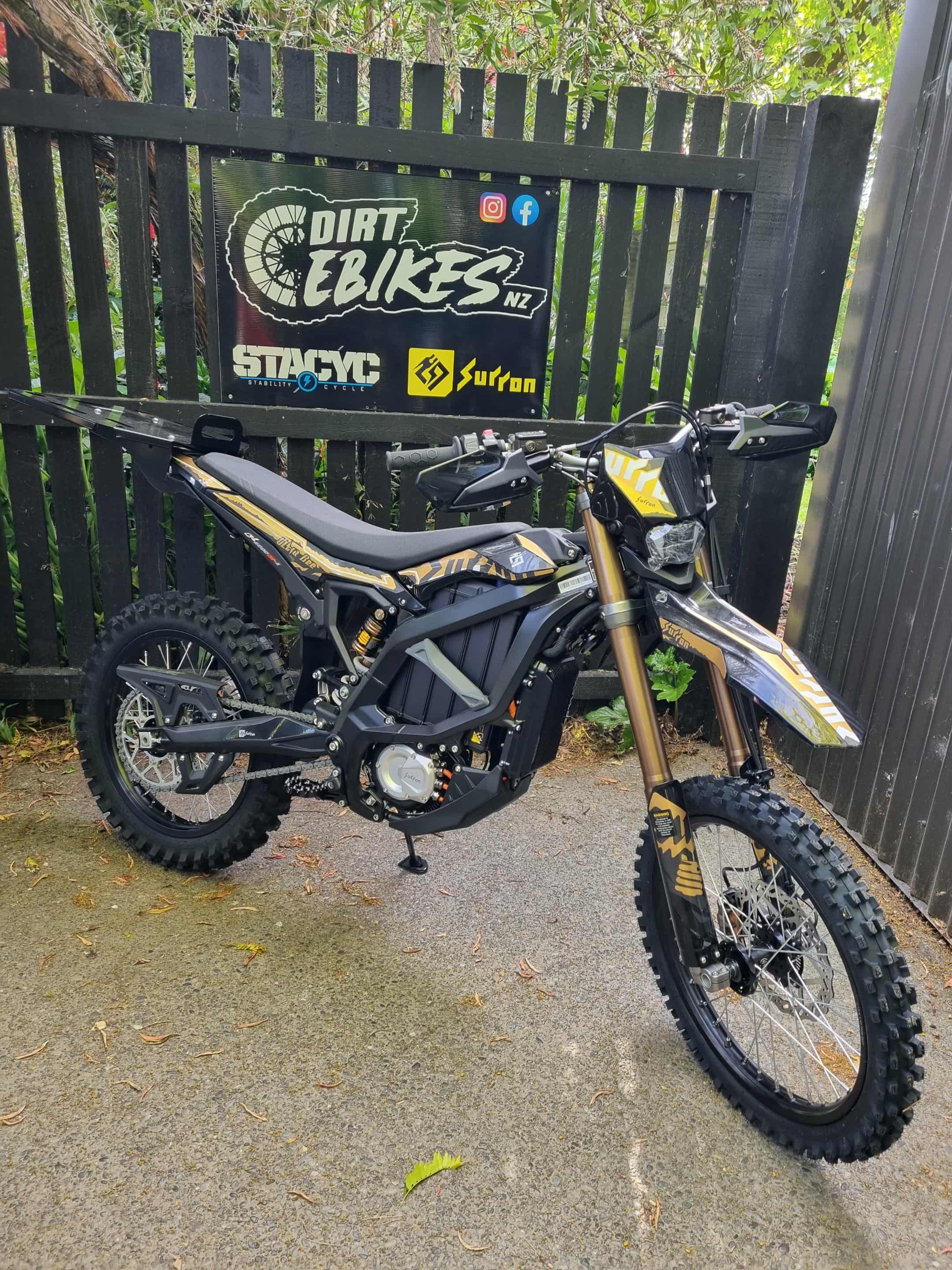 Electric dirt bike in front of a branded fence with 'Dirt E Bikes NZ' and 'Surron' logos.
