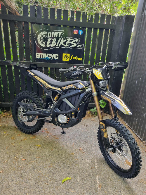 Electric dirt bike in front of a branded fence with 'Dirt E Bikes NZ' and 'Surron' logos.